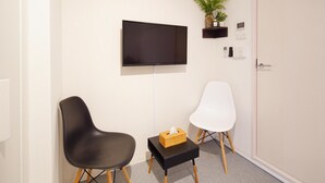 Ett rum med en platt-TV som är monterad på väggen, två stolar, ett litet bord med en näsdukslåda och en krukväxt på en hylla.