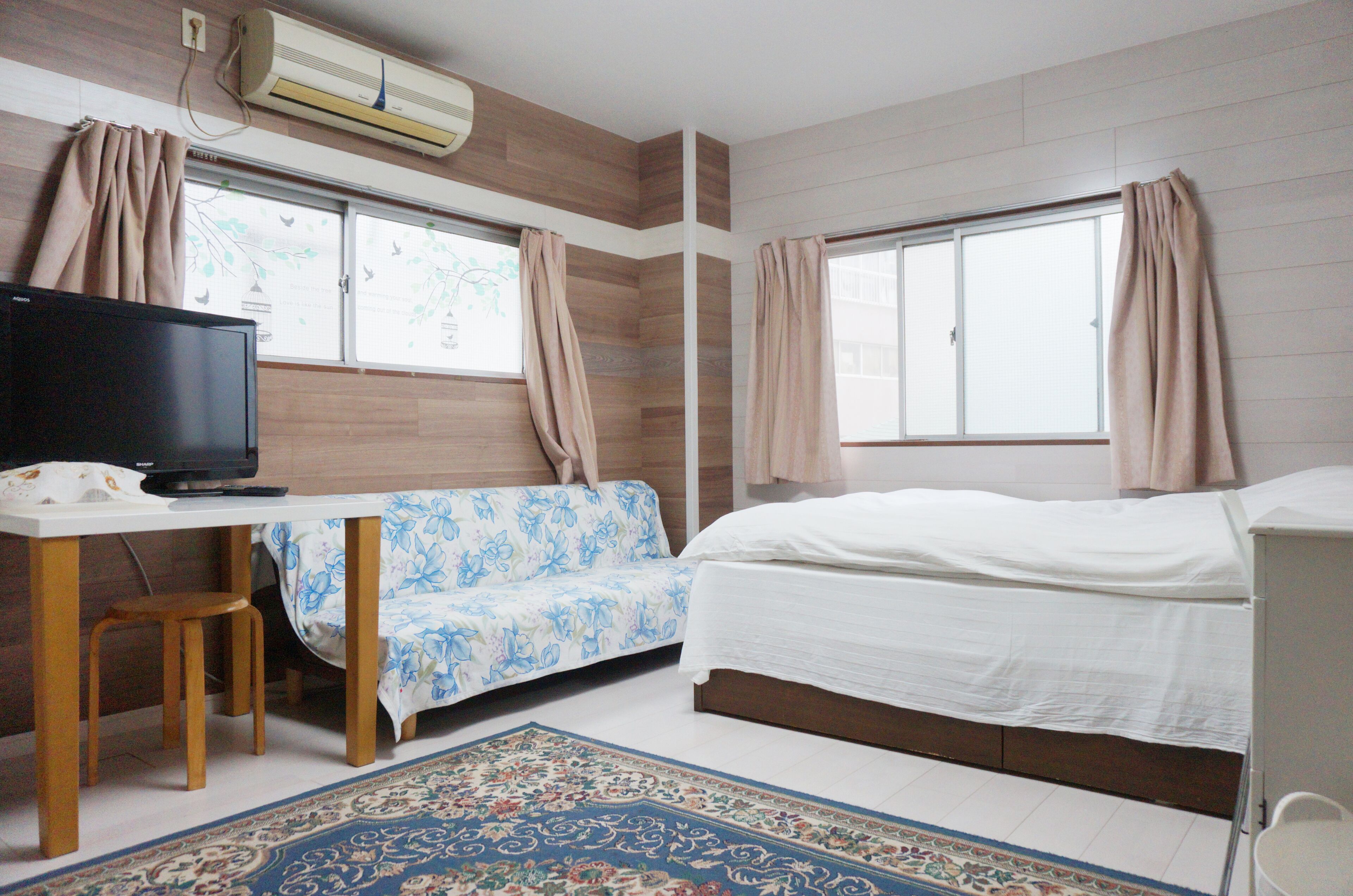 A room with a bed, a sofa, a desk with a television, and an air conditioning unit.