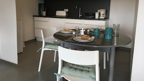 Cozinha moderna com área de refeições. Inclui micro-ondas, torradeira e cafeteira na bancada. Há duas cadeiras com almofadas e uma mesa de jantar redonda posta com pratos, copos e uma tigela.