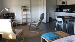 Uma sala moderna com televisão, uma cadeira de madeira, um sofá, uma mesa de jantar com cadeiras, uma área de cozinha com micro-ondas e um lustre grande.