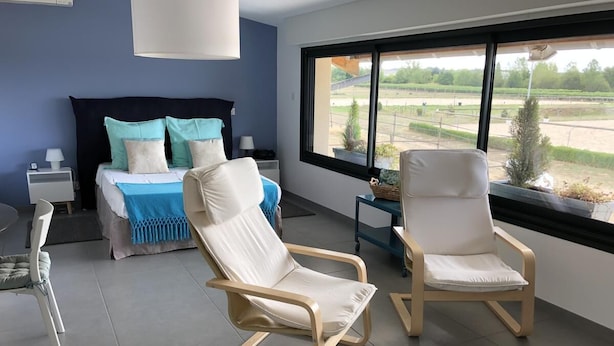 Quarto moderno com uma cama, duas poltronas brancas, uma mesinha de cabeceira e uma janela com vista para um campo.