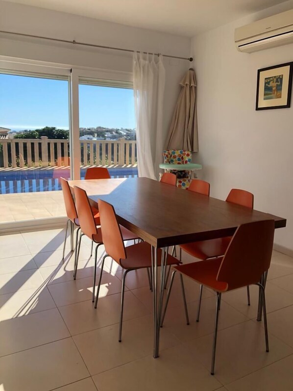 A dining area with a large wooden table and orange chairs, a small table with a colorful polka-dotted tablecloth, and a view of a pool and white fence through large windows.