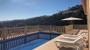 A swimming pool with lounge chairs and an umbrella, surrounded by a wooden fence, overlooking a hillside with buildings. - Villa Zoe - 4 bed - sleeps 8 - sea views - BOOK NOW!!!! (Benitachell, Alicante)
