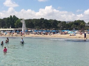 A beach with people swimming and sunbathing, a boat, and a row of blue umbrellas.