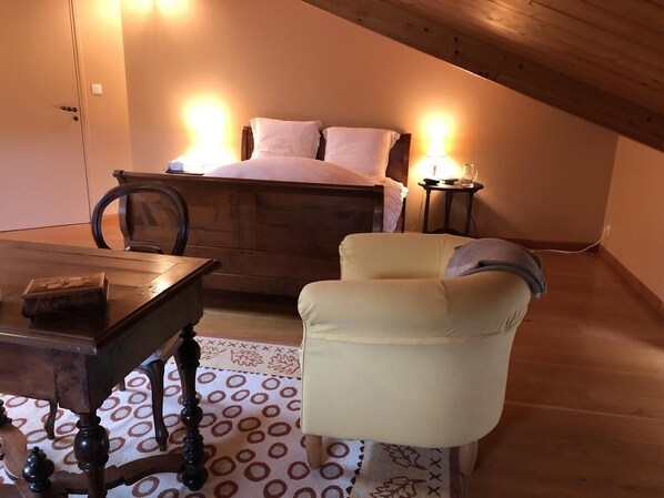 A bedroom with a wooden bed, a small table, a chair, and a bedside table with a lamp. - Vaste chambre toulousaine 18ème proche Toulouse (Bouloc)