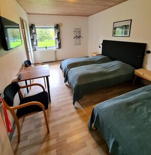 A room with two beds, a table, and a chair. There is a window with curtains, a radiator, and a picture on the wall. - Nordgården Pension (Samsø)