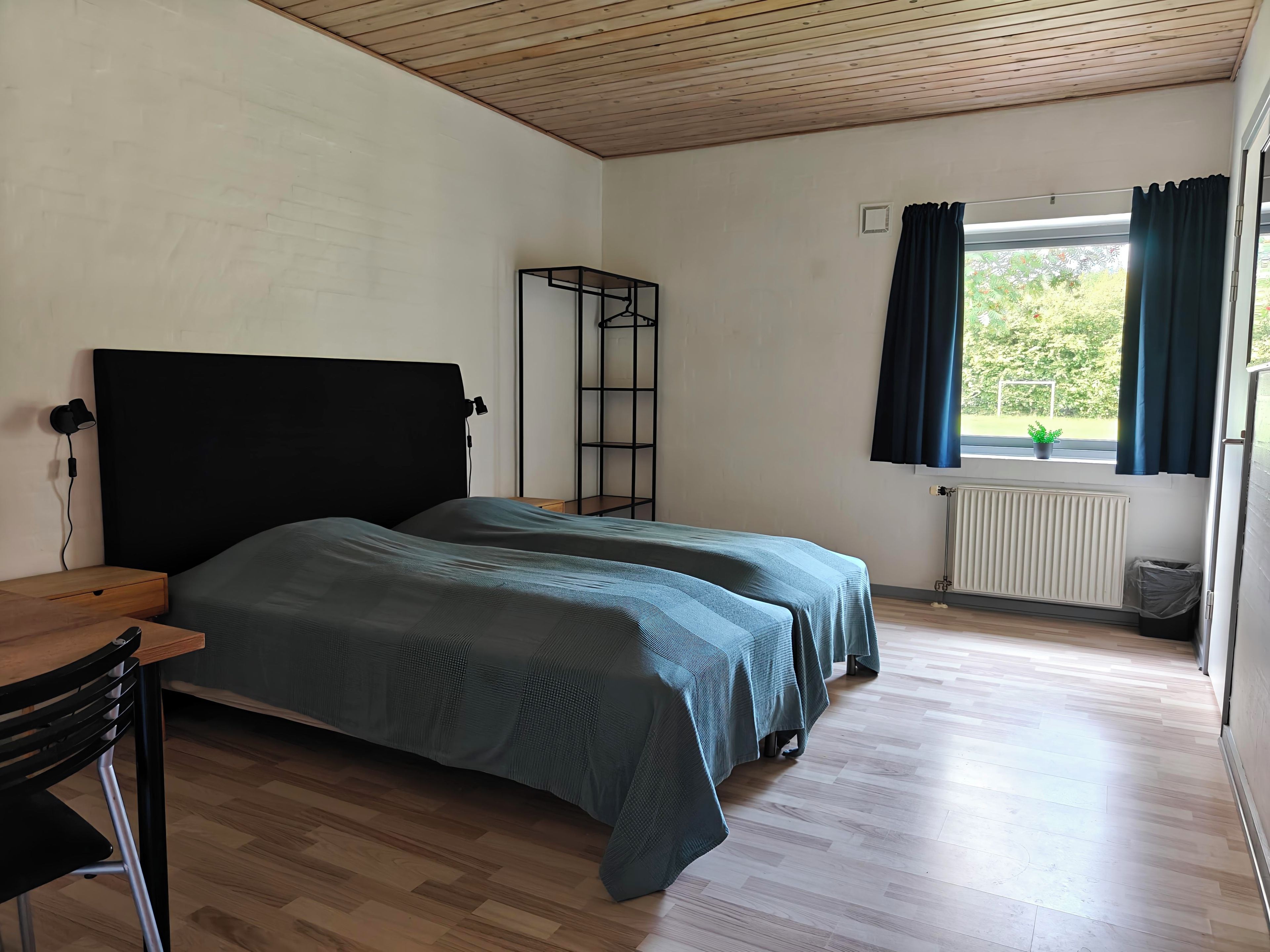 A room with a bed, a desk, a chair, a radiator, and a window with curtains.