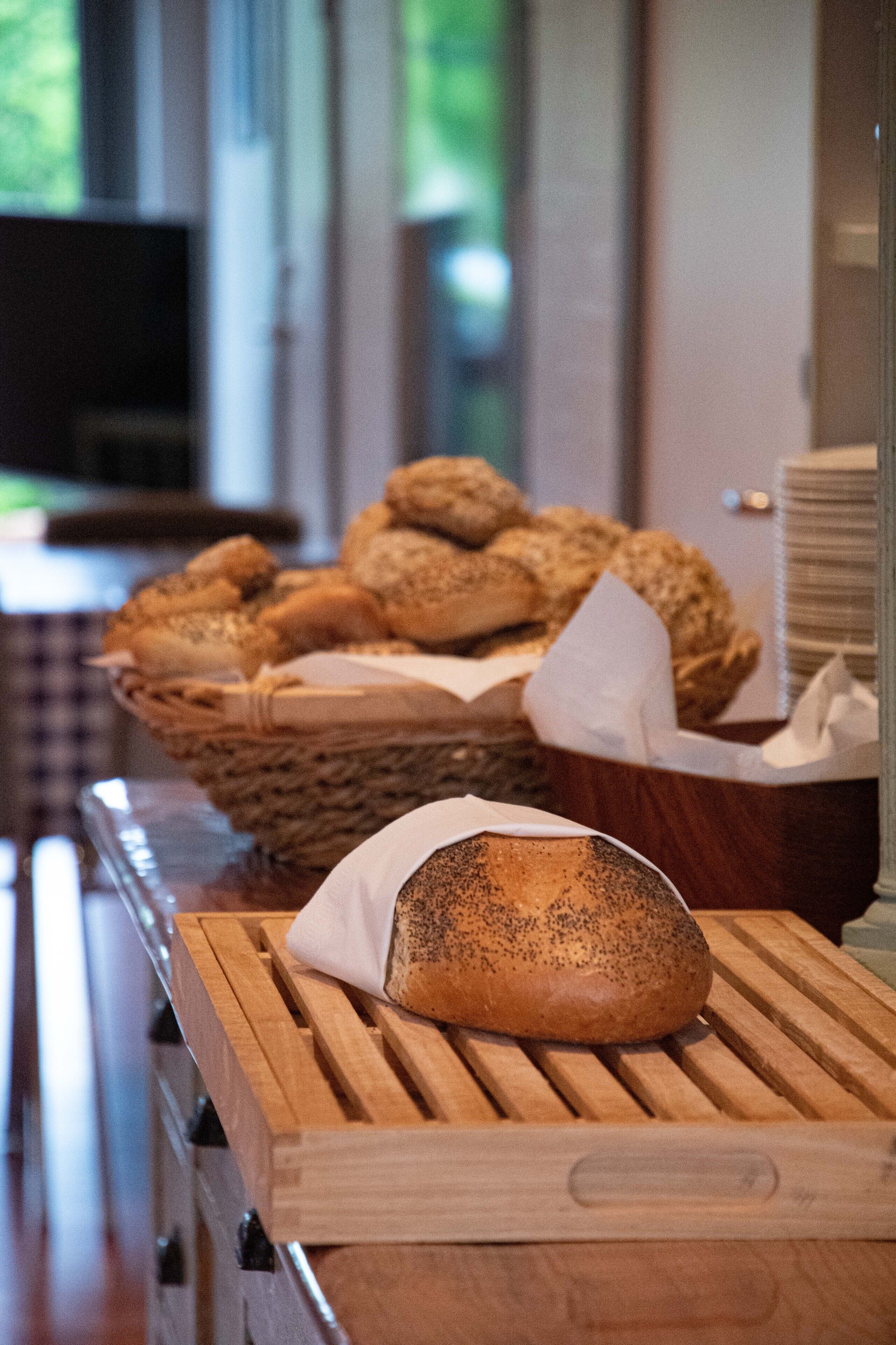 Una exhibición de pan de madera con una hogaza sobre una tabla de cortar, una cesta de panes variados y una pila de platos al fondo.