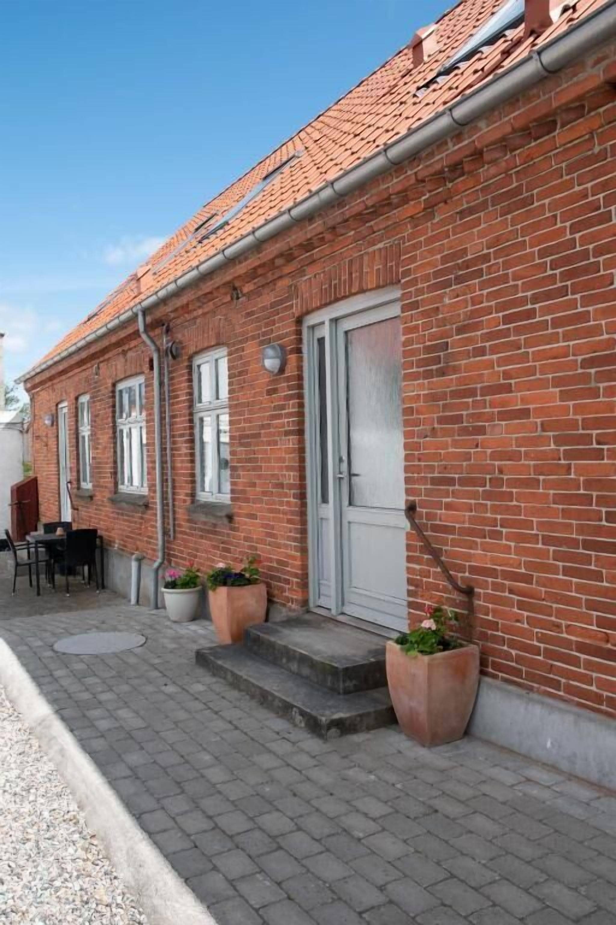Ein rot backenfassendes Gebäude mit einem Ziegeldach, einer weißen Tür und Stufen zum Eingang. Vor der Fassade befinden sich Kübelpflanzen und ein Tisch mit Stühlen.