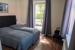 A bedroom with two beds, a window with curtains, a radiator, and a chair. - Nordgården Pension (Samsø)