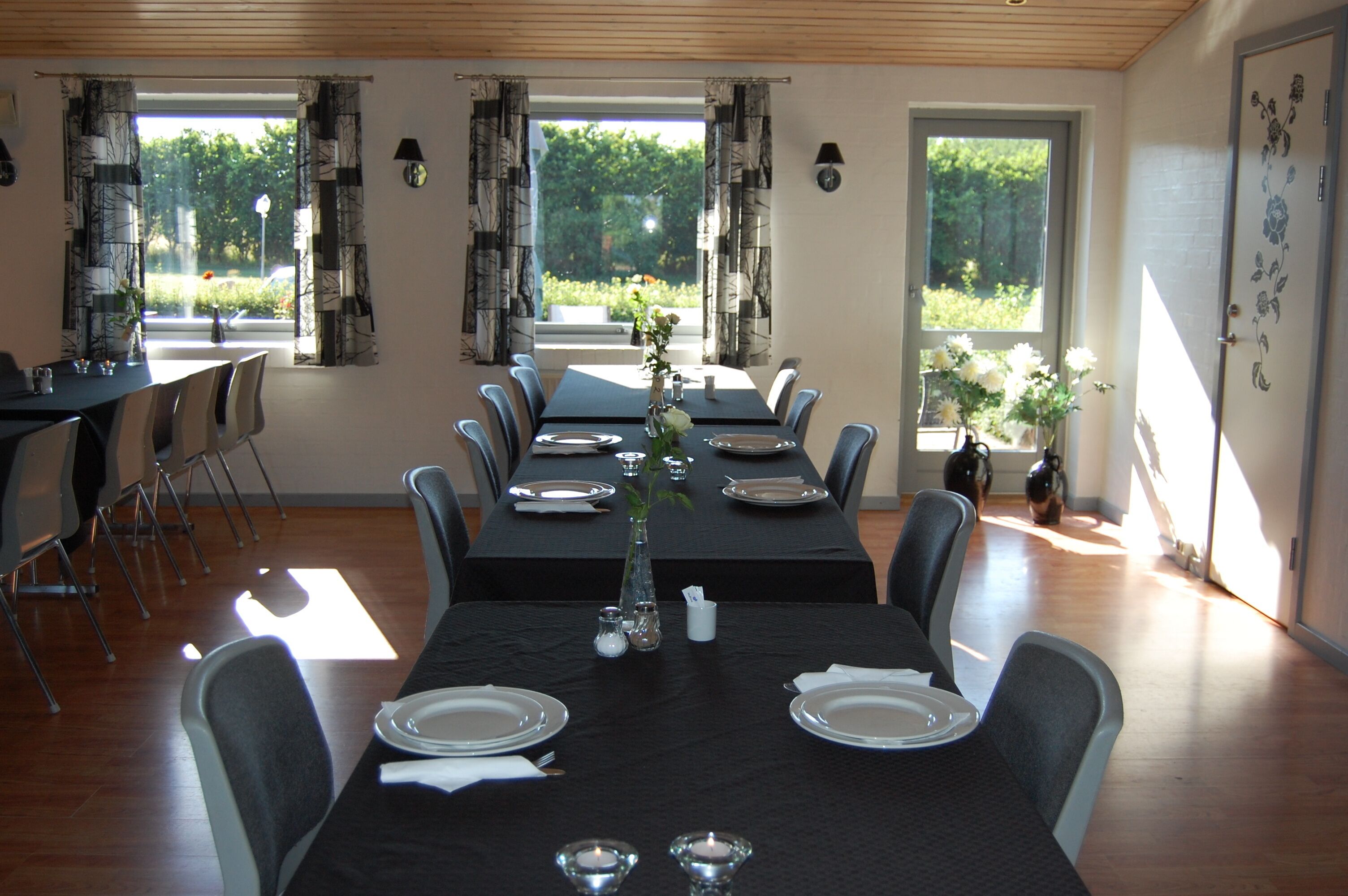 A dining room with a long table set for a meal, surrounded by chairs, with windows offering a view of greenery outside.