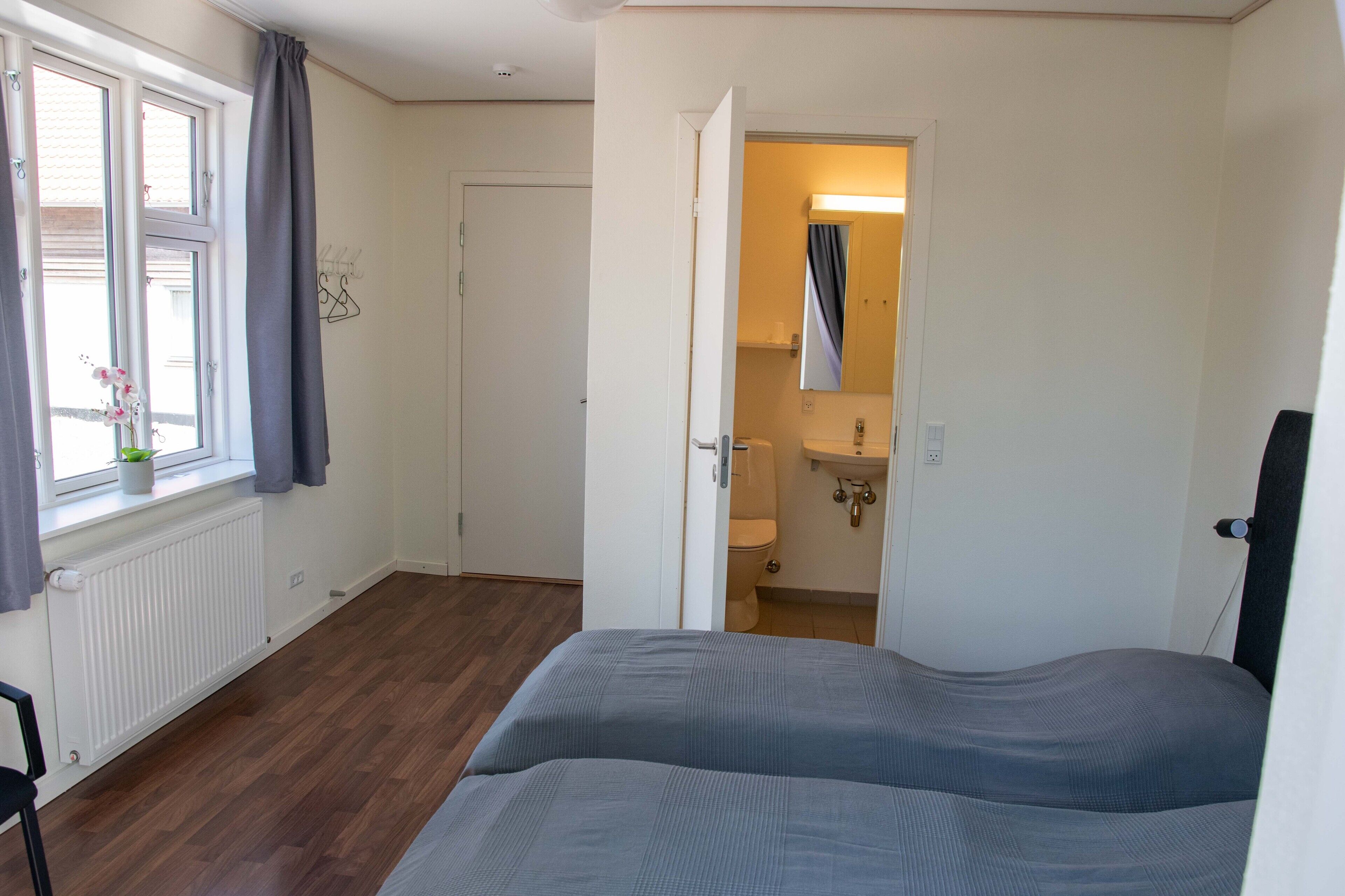 Een slaapkamer met een bed, een raam met gordijnen, een radiator en een deur naar een badkamer.
