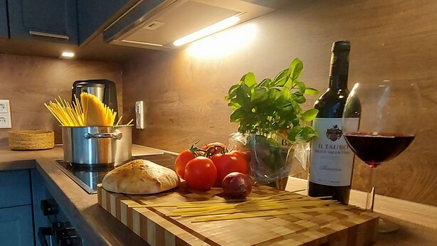 A kitchen counter with a cutting board, a bottle of wine, a glass of wine, a plant, tomatoes, a loaf of bread, and a pot of spaghetti. - Tante Maaß – maisonette flat, shower, washing machine, parking space, TV, WiFi (Flensburg)