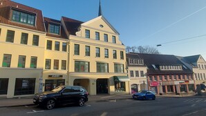 A street view with a building featuring a shop named 'Academy' on the ground floor, a church steeple, and a blue car parked on the street. - Tante Maaß – maisonette flat, shower, washing machine, parking space, TV, WiFi (Flensburg)