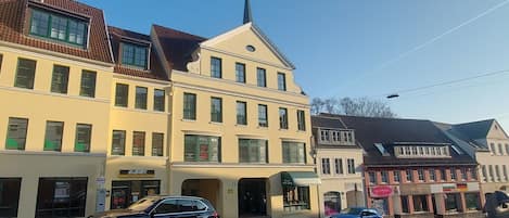 A street view with a building featuring a shop named 'Academy' on the ground floor, a church steeple, and a blue car parked on the street.