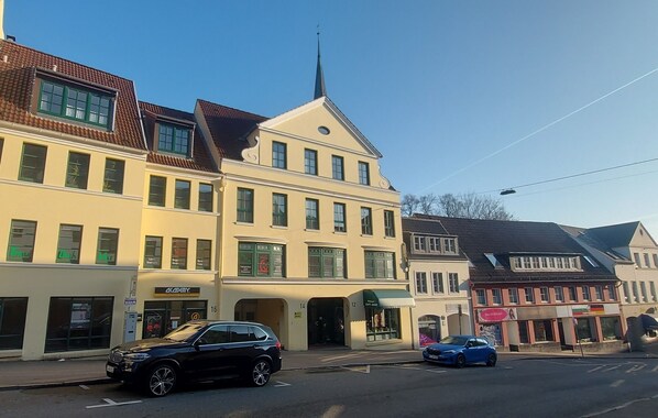 A street view with a building featuring a shop named 'Academy' on the ground floor, a church steeple, and a blue car parked on the street.
