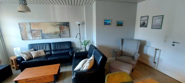 Een woonkamer met een zwarte leren bank, een fauteuil met een patroon, een houten salontafel en een vloerlamp. Er hangen ingelijste foto's aan de muur en boven de bank een groot abstract schilderij.