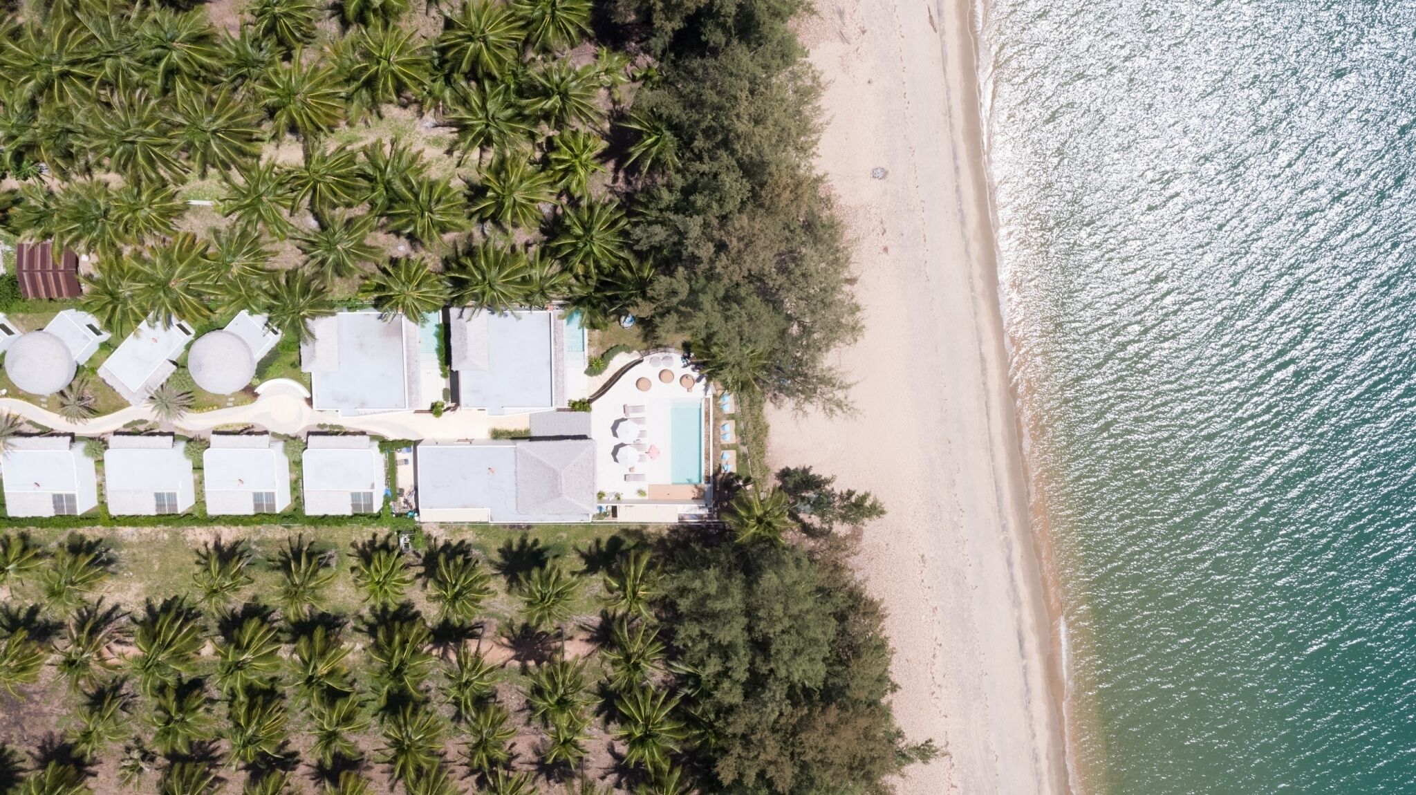 Vista aérea de un resort costero con múltiples alojamientos tipo bungalow, un área de piscina central y acceso directo a la playa.