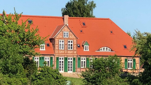 A large, two-story house with a red roof and green shutters, surrounded by trees and a grassy field. - Hirschaue - Redentin manor house (Krusenhagen)