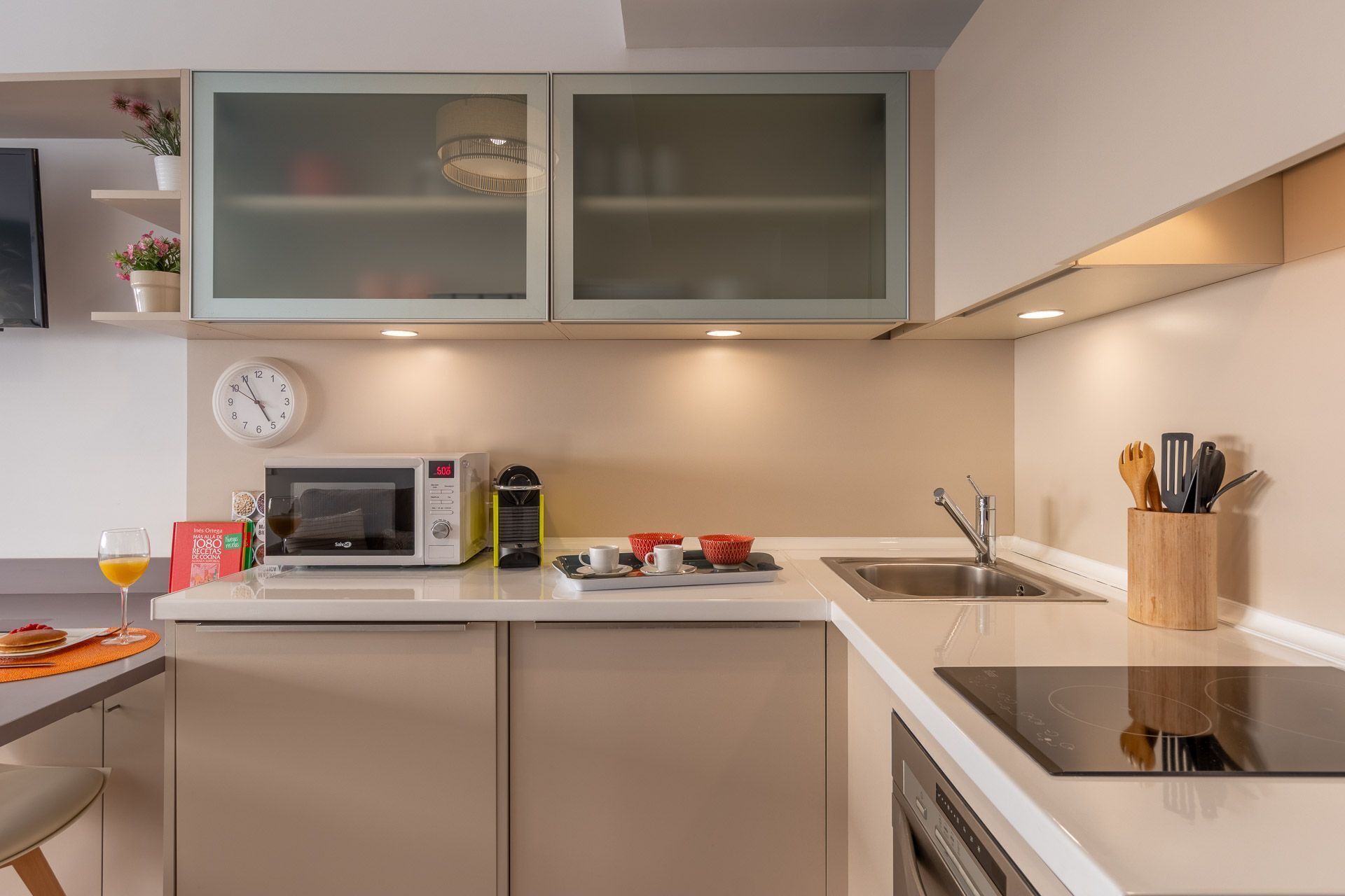 A modern kitchen with a microwave, toaster, and sink. There is a clock on the wall and a potted plant on the shelf.