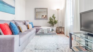 Uma sala de estar moderna com um sofá cinzento, almofadas coloridas, uma mesa de centro de vidro, uma televisão de tela plana grande e um abajur.