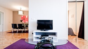 Un salon moderne avec un téléviseur à écran plat posé sur un meuble blanc, une table basse en verre et un tapis violet. On aperçoit à l’arrière-plan un coin repas avec une table et des chaises.