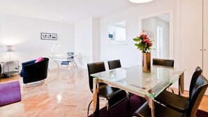 Un espace repas moderne avec une table et des chaises en verre, un tapis violet, un bureau avec une lampe, et une vue sur une cuisine et une salle de bain à l’arrière-plan.