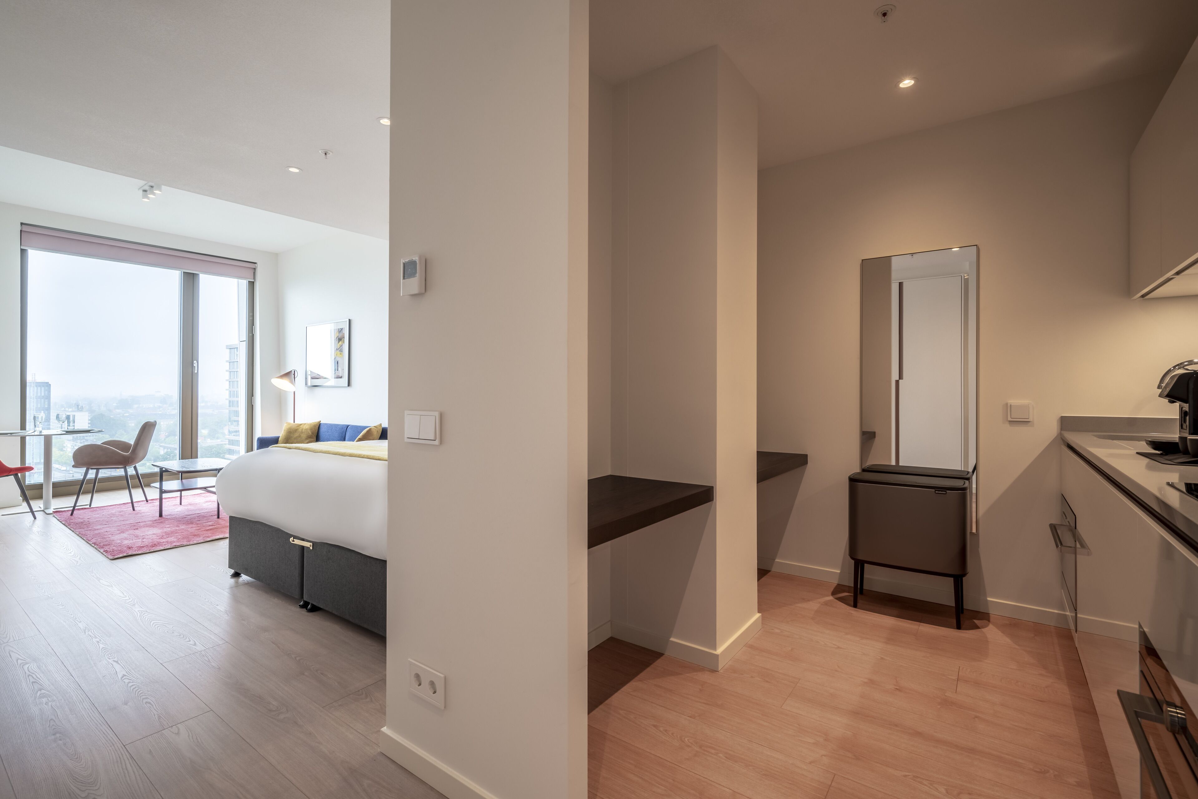 A modern hotel room with a large bed, a desk, a small bathroom, and a view of the city.