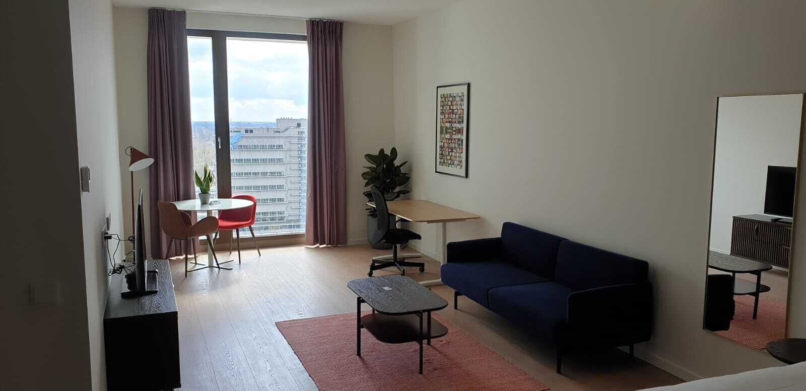 Una sala moderna con un sofá, una mesa de comedor con sillas, una pequeña mesa de centro, un televisor y un ventanal con cortinas.