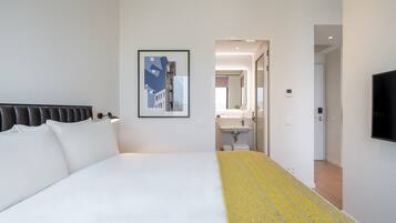 A modern hotel room with a large bed, a framed picture on the wall, a bathroom visible through an open door, and a TV mounted on the wall.