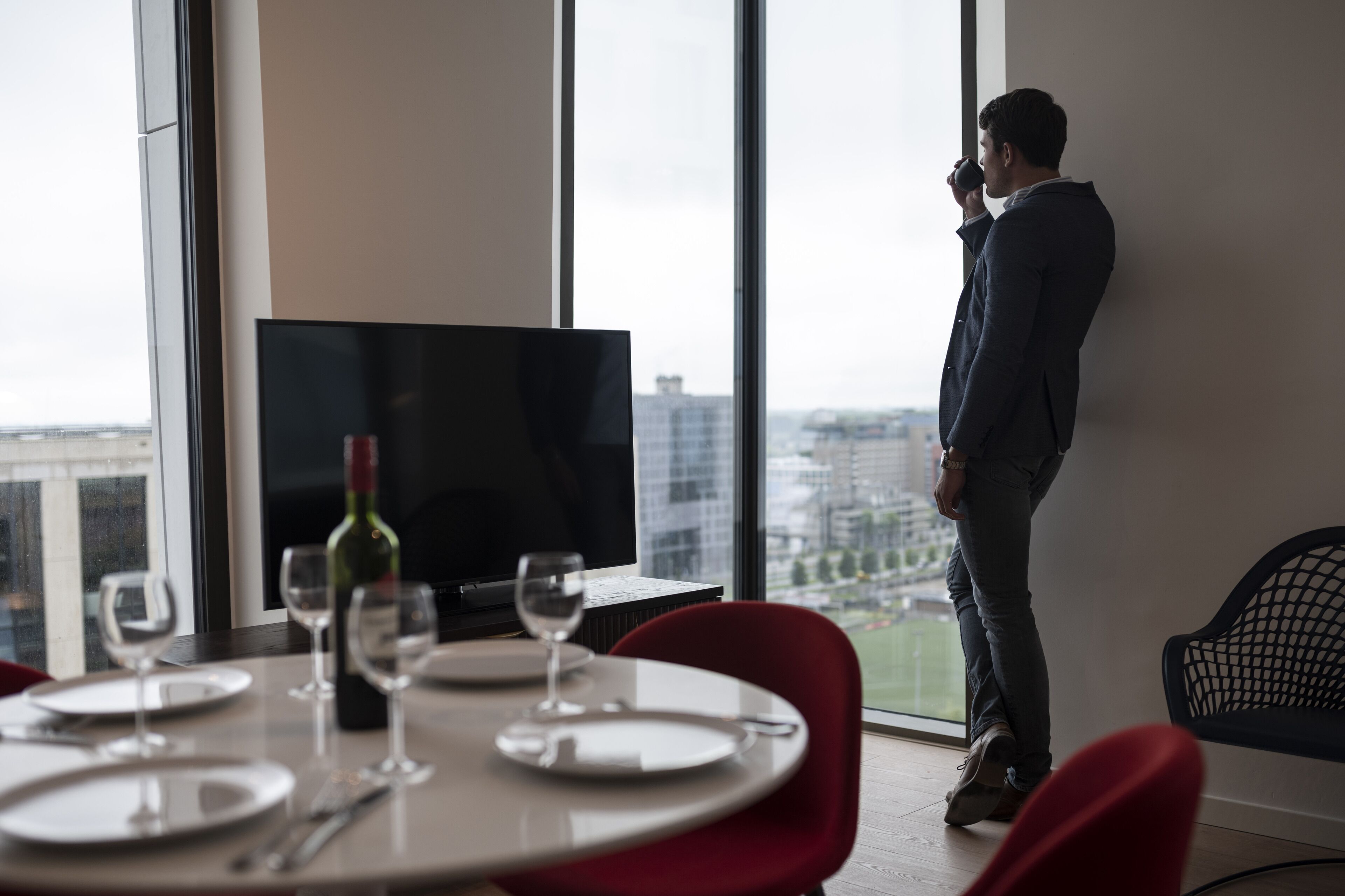 Een moderne hotelkamer met een eettafel gedekt voor twee personen, een groot raam met uitzicht over de stad en een flatscreen televisie op een standaard.