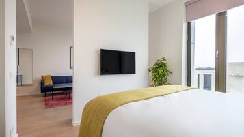 A modern hotel room with a large bed, a flat-screen TV mounted on the wall, a sofa in the background, and a plant by the window.
