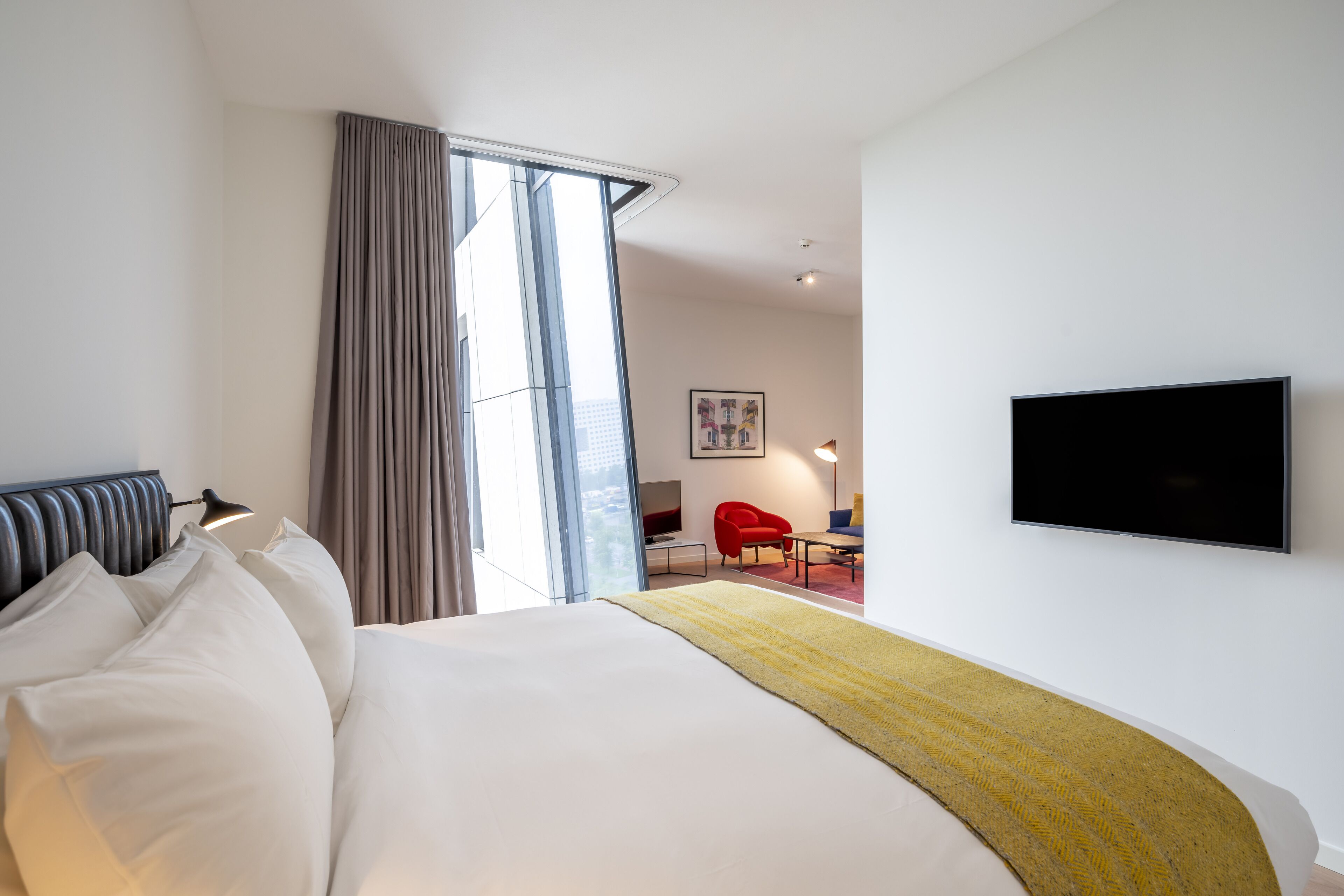 A modern hotel room with a large bed, a flat-screen TV, a red chair, and a large window with curtains.