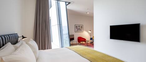 A modern hotel room with a large bed, a flat-screen TV, a red chair, and a large window with curtains.