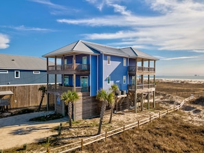 A two-story beachfront house with balconies, a wooden deck, and a sandy beach in the background.