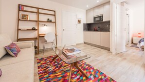 Un espace de vie compact comprenant un canapé, une table basse en verre, un tapis coloré, une chaise blanche, une bibliothèque et un coin cuisine avec un four à micro-ondes, ainsi qu’une porte donnant sur une autre pièce.