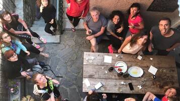 Eine Gruppe von Personen sitzt draußen an einem Holztisch, im Hintergrund eine Steinmauer und eine Tafel.