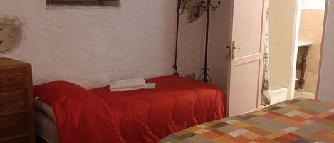 A bedroom with two beds, a window with green shutters, a door with a mirror, and a small table.