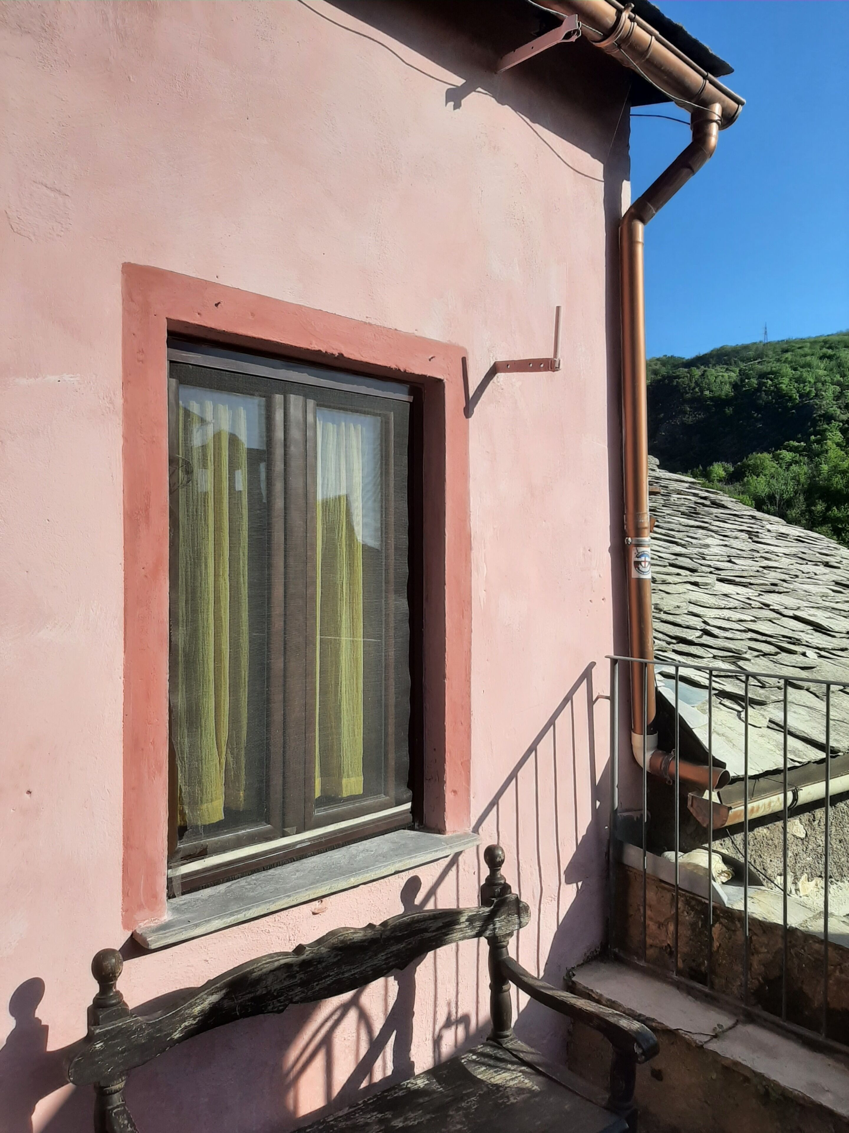 Un edificio rosa con ventana, barandilla metálica y vistas a un tejado de piedra y vegetación.