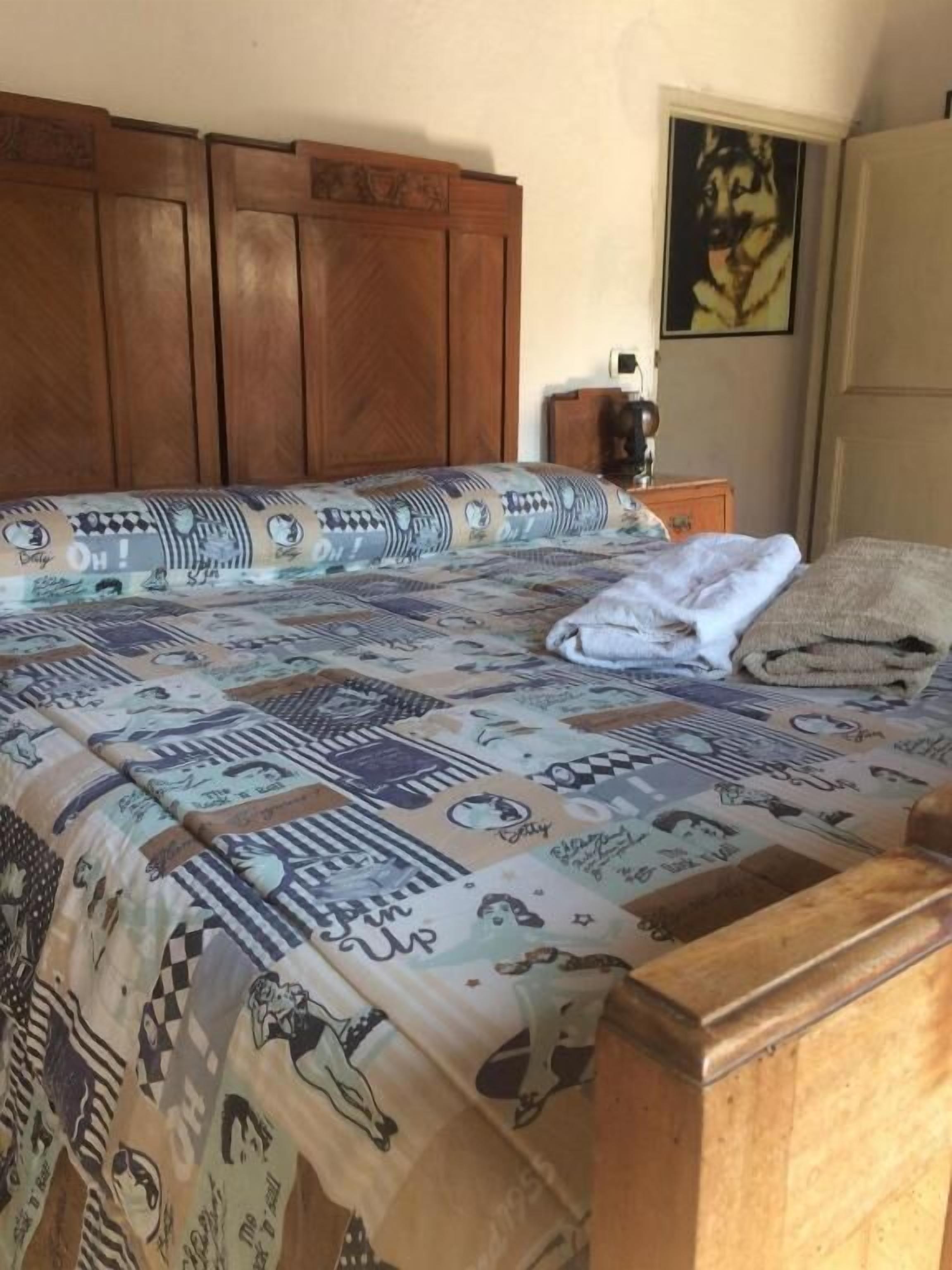 Una cama con una colcha estampada, un cabecero de madera, una mesita de noche con una lámpara y un cuadro enmarcado en la pared.