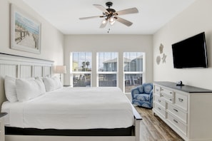 A bedroom with a bed, a ceiling fan, a TV, a dresser, and a window with a view of the outside.