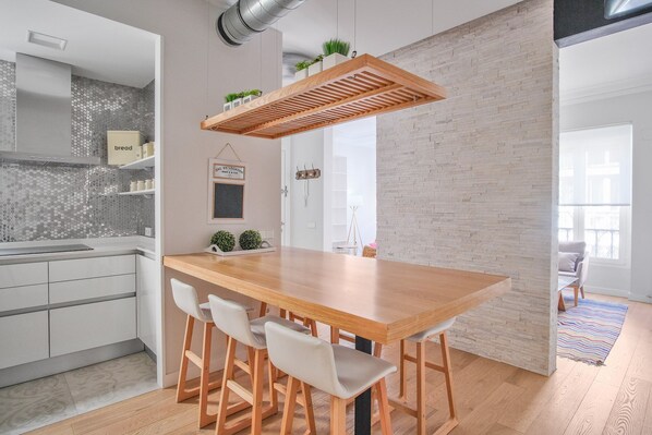 A modern kitchen with a wooden dining table and bar stools, a hanging light fixture, and a brick wall.