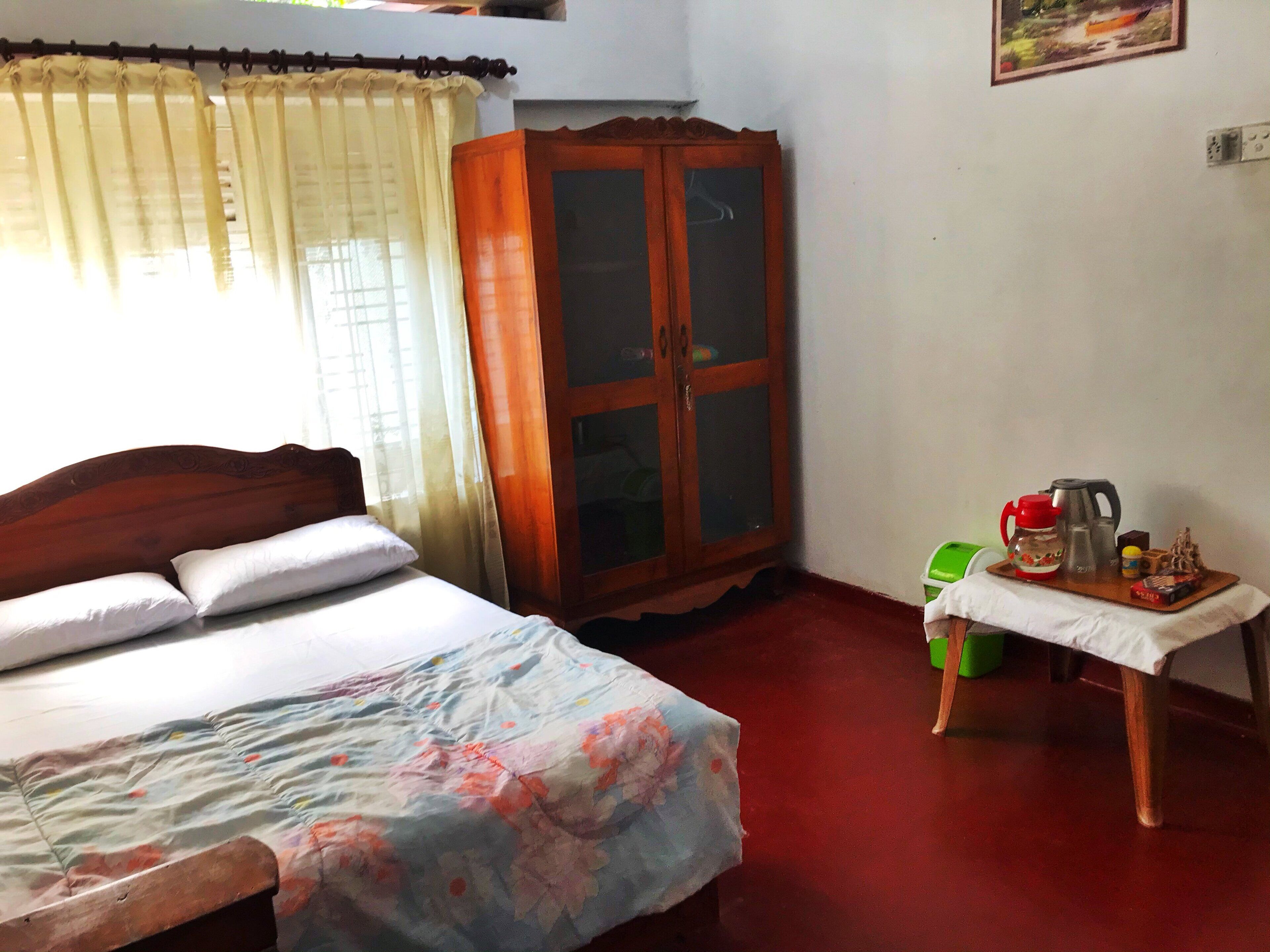 Quarto com cama, um armário de madeira, uma mesinha com chaleira e uma cadeira verde.