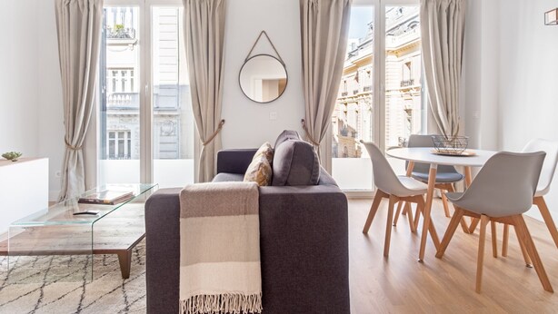 Un salón moderno con un sofá, una mesa de comedor con sillas, ventanas grandes con cortinas y vistas a edificios.