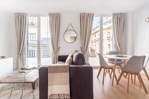 Een moderne woonkamer met een bank, een eettafel met stoelen, grote ramen met gordijnen en uitzicht op gebouwen.