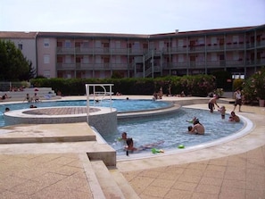 A swimming pool area with multiple pools, a diving board, and a building with balconies in the background.