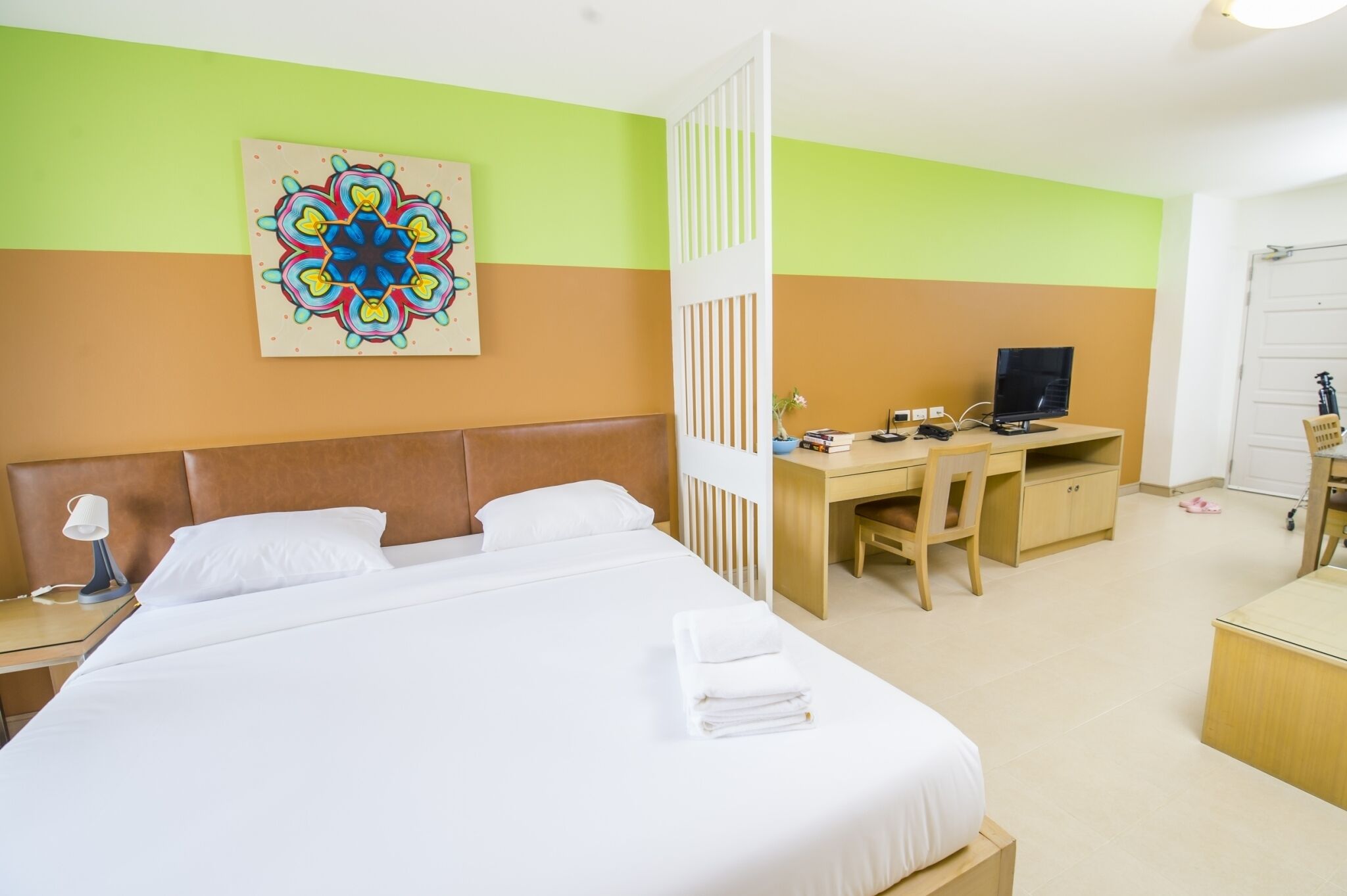 A hotel room with a large bed, a desk with a computer, a TV, and a colorful artwork on the wall.