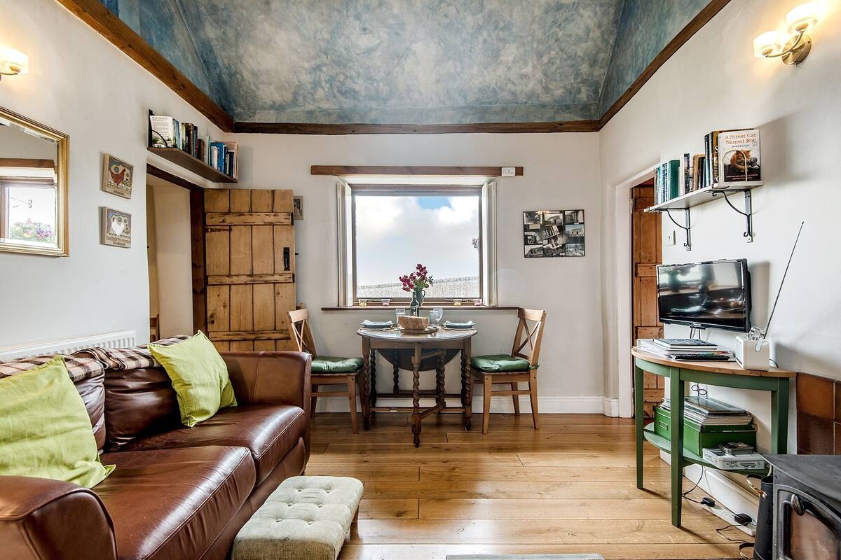 Una sala acogedora con un sofá de cuero marrón, una mesa de comedor de madera con sillas, un televisor sobre un mueble bajo y una estantería.