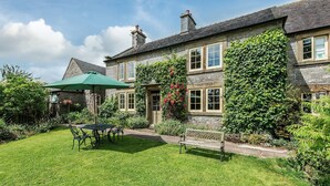 Une maison en pierre avec un jardin bien entretenu, une terrasse avec une table et des chaises, et un parasol vert.
