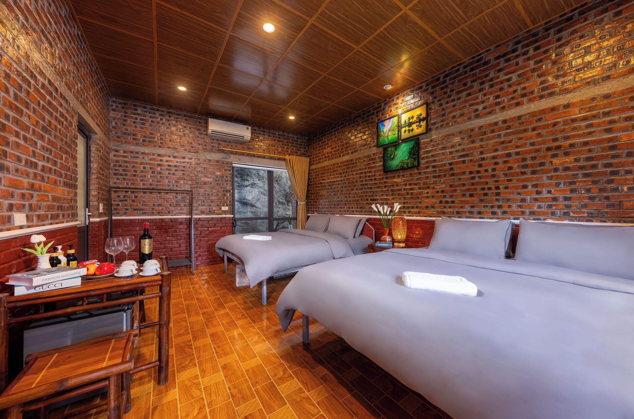 Quarto com parede de tijolos, teto de madeira, duas camas, uma mesa com objetos e uma janela.